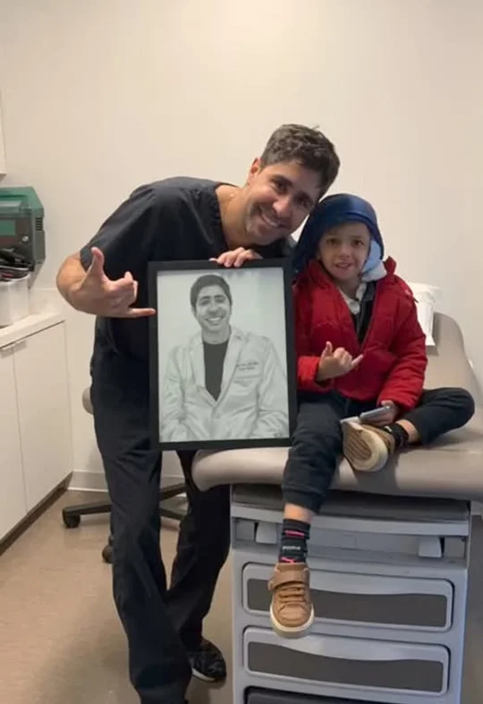 Dr. Tahiri holding a framed portrait beside a young microtia patient in an exam room